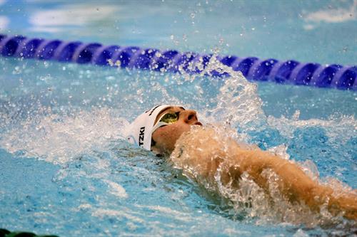 Ein Schwimmer absolviert Rücken in einem Schwimmbecken. Wasser spritzt um sein Gesicht und seine Arme, im Hintergrund ist eine Bahnbegrenzung zu sehen.